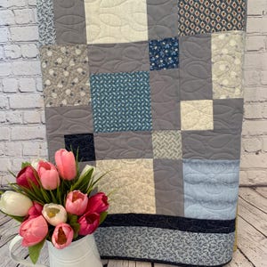 May include: A patchwork quilt in shades of grey, blue, and cream, with various patterned squares. The quilt features floral and geometric designs, with quilted details. A bouquet of pink and white tulips in a white jug sits in front of the quilt.