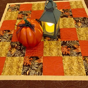 Peut inclure: Un chemin de table patchwork carré composé de blocs alternés en tissu orange à pois blancs et en tissu sombre à motifs de feuilles d'automne. Une citrouille orange vif avec un ruban à pois noirs et une lanterne grise éclairée sont posés dessus.