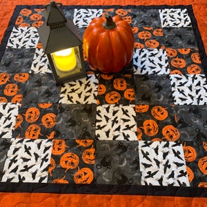 Peut inclure: Une courtepointe noire et blanche sur le thème d'Halloween. La courtepointe présente une variété de citrouilles, de chauves-souris et de chats. Il y a une lanterne noire et une grande citrouille orange au centre de la courtepointe.