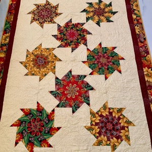 May include: A beige quilt with a repeating pattern of nine star-shaped fabric blocks. Each block features a different kaleidoscope-like design with shades of red, green, brown, and yellow.