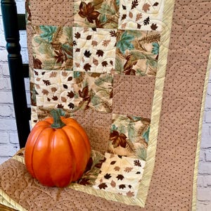May include: An orange pumpkin sits on a quilted blanket with a fall leaf pattern. The blanket features squares of fabric with leaves in shades of brown, green, and cream. The blanket is trimmed with a light yellow border and a brown dotted pattern.