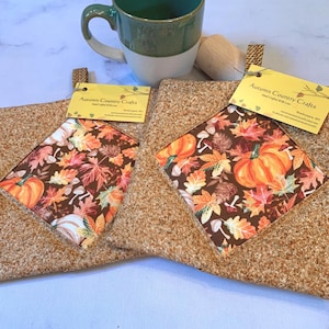 May include: Two square fabric pot holders with a brown textured border and a central panel featuring a pattern of orange pumpkins, brown pinecones, and colorful autumn leaves. Each pot holder has a yellow tag labeled Autumn Country Crafts.