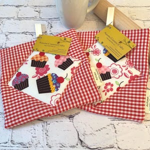 May include: Two quilted pot holders with a red and white gingham pattern border. The center features a white fabric with cupcake and floral designs. Each pot holder has a tag that reads "Autumn Country Crafts".