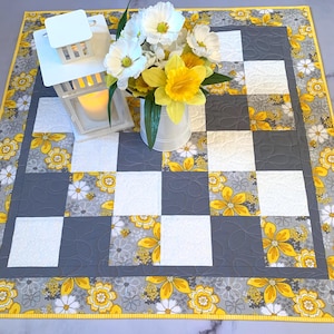 May include: A quilted table runner with a checkerboard pattern of gray and white squares, accented with yellow floral designs. A white lantern with a lit candle and a bouquet of white and yellow flowers in a white bucket sit on the runner.