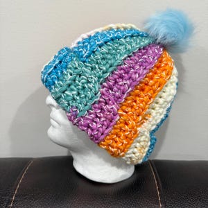 May include: A colorful, striped crochet beanie with a light blue pom-pom. The hat features bands of turquoise, purple, orange, and white, with a white thread woven throughout. The beanie is displayed on a white mannequin head.