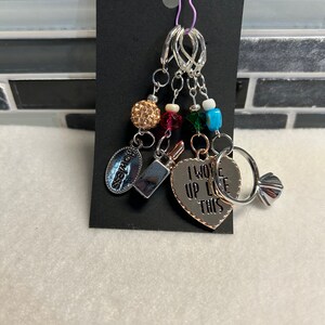 May include: A collection of silver charms and beads, including a lipstick, a heart with the text "I WOKE UP LIKE THIS", and a charm that says "flawless". The charms are attached to a black tag with a purple safety pin.