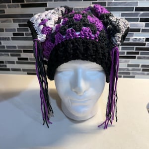 May include: A handmade crochet beanie featuring a granny square pattern in shades of purple, black, and grey. The hat has a solid black ribbed brim and long, dangling yarn tassels in purple and black hanging from both sides of the head covering.