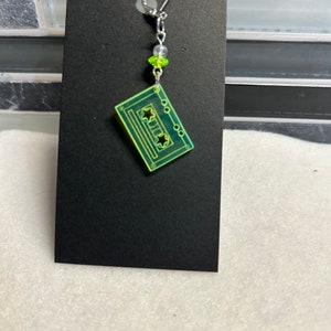 May include: A single earring featuring a neon green cassette tape design. The earring hangs from a silver-colored hook and chain, with clear and green beads. The cassette tape has star-shaped cutouts and etched details.