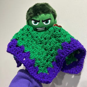 May include: A plush Hulk doll with a green head and a green and purple crocheted poncho. The Hulk's face is green with black eyebrows and a stern expression. The poncho is green with purple trim.