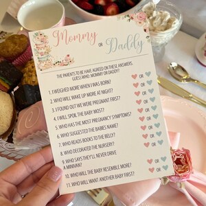 May include: A baby shower game card with the text "Mommy or Daddy" in pink and blue script. The card features questions about the parents, with pink and blue heart check boxes. Floral and cupcake illustrations decorate the card.