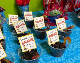 May include: A table setting with a blue tablecloth and a variety of party decorations. There are several clear plastic cups filled with chocolate pudding, gummy worms, and Oreo cookie crumbs. Each cup has a small sign that reads "There's a snake in my boot!"