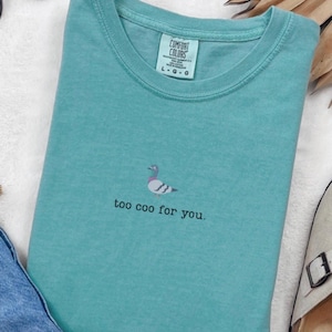 May include: A teal Comfort Colors t-shirt with a small embroidered pigeon design and the phrase "too coo for you." The shirt has a crew neck and a tag in the upper center.