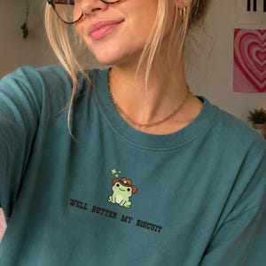May include: Teal t-shirt featuring a cartoon frog wearing a cowboy hat and the phrase "Well Butter My Biscuit." The frog is green with a brown hat and yellow stars. The shirt has a crew neck and short sleeves.