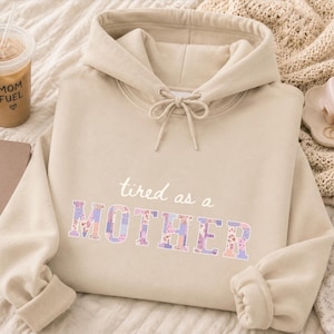 Può includere: Una felpa con cappuccio beige con la scritta "tired as a MOTHER" in bianco e lettere a fantasia floreale. La felpa ha un cappuccio con coulisse e polsini a coste. È visibile una tazza di caffè con "MOM FUEL".