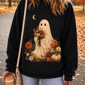 May include: A black long-sleeve sweatshirt featuring a graphic print of a white ghost holding a bouquet of autumn flowers. The design includes a crescent moon above the ghost and a pumpkin surrounded by fall foliage at the base of the illustration.