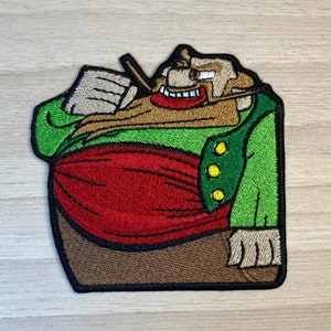 May include: Embroidered patch of a cartoon character with a large belly, wearing a green jacket with yellow buttons, a red sash, and a brown beard. The character is holding a pipe in his right hand.