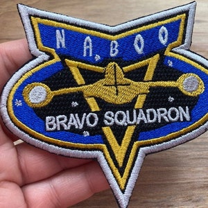以下が含まれることがあります： 「NABOO BRAVO SQUADRON」という文字と、スタイリッシュな宇宙船のデザインが施された、青と金の刺繍パッチです。