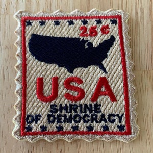 May include: A beige fabric patch with a red border and a red and blue embroidered image of the United States. The patch has the text "USA" in red and "SHRINE OF DEMOCRACY" in blue. The patch also has a red "25¢" in the upper right corner.