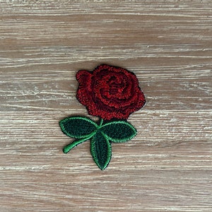 May include: An embroidered red rose with green leaves on a wooden background.