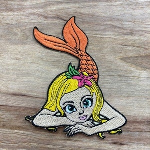 May include: Embroidered iron-on patch of a mermaid with blonde hair, a pink flower, and an orange tail. The mermaid is lying on her stomach with her arms crossed under her chin.