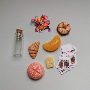Könnte beinhalten: Eine Sammlung von Miniatur-Lebensmitteln, darunter ein Croissant, ein Donut, eine Orangenscheibe, ein kleines Brot, eine Flasche und eine kleine Tüte mit bunten Streuseln.