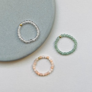 Könnte beinhalten: Drei kreisförmige Perlenringe, die jeweils mit einer kleinen goldfarbenen Metallperle akzentuiert sind. Ein Ring besteht aus klaren, durchsichtigen Perlen, einer aus hellgrünen Perlen und einer aus blass pfirsichfarbenen Perlen.