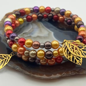 May include: A multi-strand bracelet featuring colorful beads in shades of red, orange, yellow, and purple. The bracelet is adorned with gold-colored leaf charms. The jewelry is displayed on a decorative stone surface.