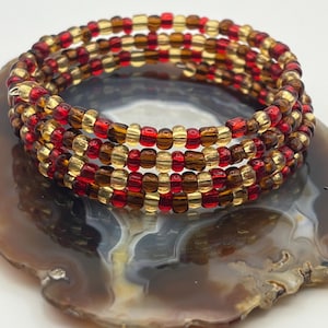 Könnte beinhalten: Ein Wickelarmband mit roten, goldenen und braunen Perlen. Das Armband besteht aus mehreren Strängen kleiner, runder Perlen, wodurch ein geschichteter Effekt entsteht. Die Perlen sind in einem sich wiederholenden Farbmuster angeordnet.