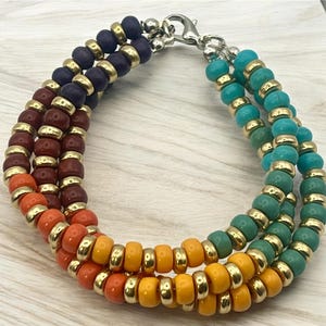 May include: A multi-strand bracelet featuring a variety of colorful beads. The bracelet has strands of navy, burgundy, orange, yellow, turquoise, and green beads, separated by gold-colored spacers. It is secured with a silver clasp.