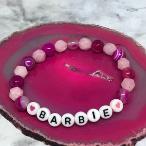 Könnte beinhalten: Ein Armband mit dem Namen "BARBIE" aus weißen Buchstabenperlen. Das Armband hat rosa und lila Edelsteinperlen. Das Armband wird auf einer rosa und weißen Achatscheibe präsentiert.