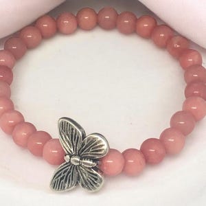 Könnte beinhalten: Ein rosafarbenes Armband mit einem silbernen Schmetterlingsanhänger. Die Perlen sind aus Rosenquarz und auf einem Gummiband gefädelt.
