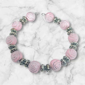 Könnte beinhalten: Eine rosafarbene Perlenkette mit silbernen Akzenten. Die Perlen sind rund und haben ein Spiralmuster. Die Kette ist hinten offen und hat einen Verschluss.