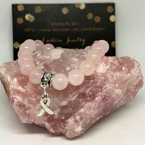 Könnte beinhalten: Ein rosafarbenes Rosenquarz-Armband mit einem silbernen Anhänger in Form eines Bandes. Das Armband liegt auf einem rosafarbenen Rosenquarz-Stein.