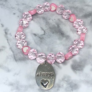 Könnte beinhalten: Rosa Kristallperlen-Armband mit einem silbernen Anhänger, auf dem "always" mit einem kleinen Herz steht.