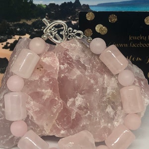 Könnte beinhalten: Ein Armband aus rosa Rosenquarz mit einem silbernen herzförmigen Verschluss. Das Armband besteht aus glatten, polierten Steinen und wird auf einem Stein präsentiert.