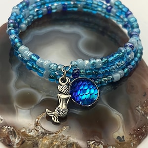 Puede incluir: Una pulsera enrollada de cuentas azules, con un dije de sirena y un dije redondo con un patrón de escamas azules. La pulsera está hecha de múltiples hilos de pequeñas cuentas.