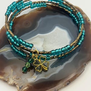 May include: A multi-strand bracelet with teal beads and gold-colored wire. A gold-colored four-leaf clover charm and a green crystal dangle from the bracelet. The bracelet is displayed on a gray and brown stone surface.