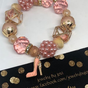 Könnte beinhalten: Ein rosa Perlenarmband mit einem High-Heel-Schuh-Anhänger. Das Armband hat verschiedene Perlenformen, darunter facettierte Kristalle, Kugeln und geometrische Formen, in Rosa-, Pfirsich- und Goldtönen. Der Anhänger ist ein rosa High-Heel-Schuh.