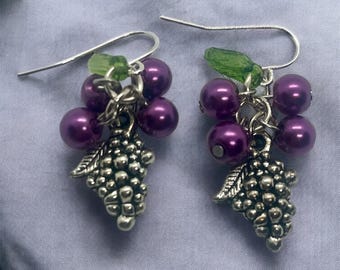 May include: A pair of drop earrings featuring silver-toned metal grape clusters, deep purple spherical beads, and small green translucent leaf-shaped accents. Each earring is suspended from a simple silver-toned metal hook for pierced ears.