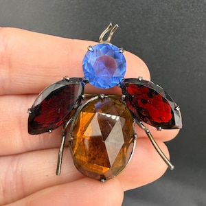 Puede incluir: Un broche con forma de insecto que presenta una piedra redonda azul facetada para la cabeza, dos piedras rojas de corte marquesa para las alas y una gran piedra ovalada de color ámbar facetada para el cuerpo, todo en un marco de metal plateado.