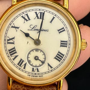 Op de afbeelding: Een goudkleurig Longines polshorloge met een crèmekleurige wijzerplaat, zwarte Romeinse cijfers en zwarte wijzers. Het horloge heeft een kleine sub-wijzerplaat voor seconden onderaan en de tekst 178 SWISS 5231 onderaan de wijzerplaat.