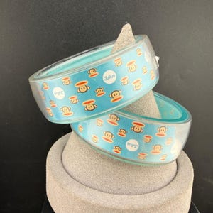 May include: Two clear, wide bangle bracelets featuring a repeating pattern of the cartoon monkey character Julius on a light blue background. Each bracelet displays the white circular Julius logo text printed on the outer surface of the clear material.