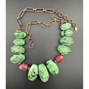 Puede incluir: Un collar con grandes piedras preciosas verdes y rojas facetadas. Las piedras están intercaladas con una cadena decorativa de color bronce texturizada. El collar tiene un cierre de gancho y una pequeña etiqueta al final de la cadena.
