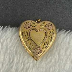 Op de afbeelding: Een hartvormig medaillon van goud met een gegraveerd bloemmotief en accenten in roségoud. Het midden van het medaillon toont een gladde, effen hartvorm omringd door een decoratieve rand, met een klein metalen lusje aan de bovenkant voor bevestiging.