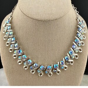 May include: A silver-tone metal necklace featuring a series of repeating ribbon-like links, each set with a round, iridescent blue and purple crystal. The necklace is constructed with a chain link design and a secure clasp mechanism at the back.
