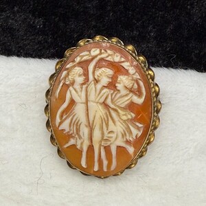 May include: An oval-shaped cameo brooch featuring a carved relief of three figures dancing in flowing dresses, set within a textured gold-toned metal frame. The carving displays cream-colored figures against a warm, translucent orange background.