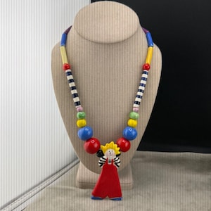 以下が含まれることがあります： 赤いドレスを着た人物のペンダントが付いたカラフルなネックレス。ネックレスは、赤、青、黄、緑、白のさまざまなビーズが特徴です。ペンダントは黄色い頭と黒い髪をしています。