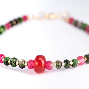 Puede incluir: Un brazalete hecho con cuentas de piedras preciosas verdes, rosas y rojas. El brazalete tiene un cierre dorado y una cuenta de piedra preciosa roja más grande en el centro.
