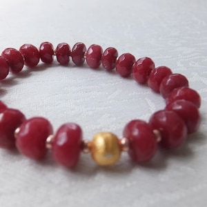 Può includere: Un braccialetto di pietre preziose rosse con un accento di perline dorate. Il braccialetto è realizzato con pietre preziose rosse sfaccettate ed è infilato su un filo sottile.