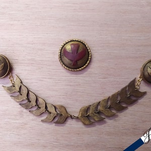 May include: A set of three gold-colored metal brooches featuring a stylized bird design. The brooches are connected by a chain of gold-colored leaves.